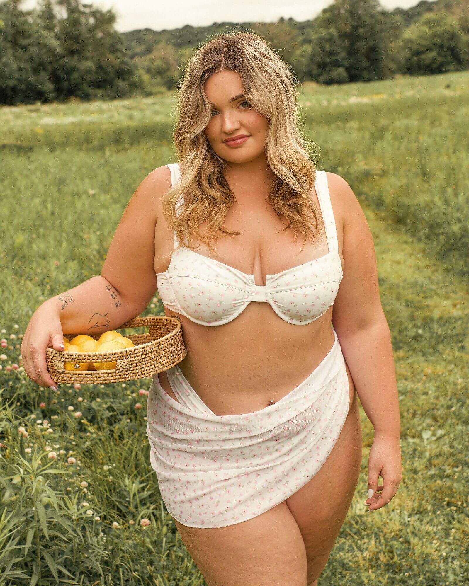 Pretty girl standing in a field and holding a basket of lemons while wearing a cute floral pink bikini along with the matching sarong.