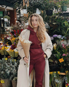 Pretty girl is holding a bouquet of flowers in front a flower shop while wearing a cute burgundy matching set and a beige trench coat.