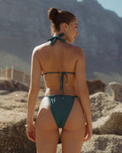 Pretty girl standing in front of rocks and looking to the right while wearing a cute shiny teal bikini.