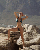 Pretty girl standing on two rocks with her hands on her head while wearing a cute shiny teal bikini.