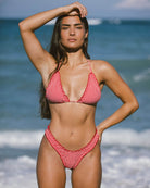 Beautiful girl standing in front of the beach with one hand on her head and the other on her hip wearing a red checkered bikini.